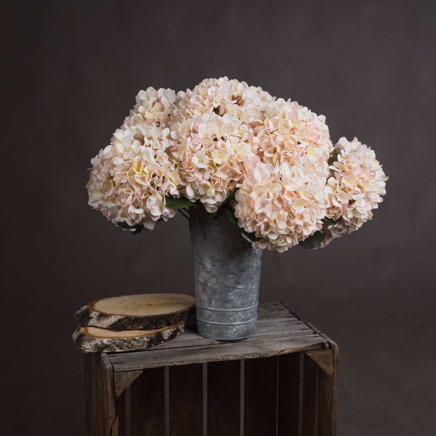 Bouquet of light pink hydrangeas in a metal vase on a wooden crate against a dark background