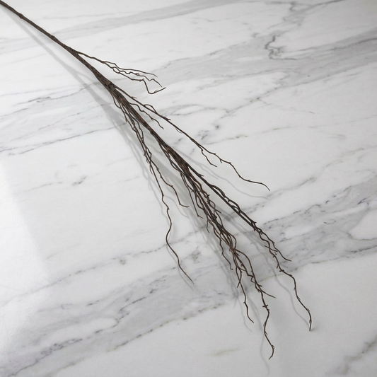 A long, thin brown twig branch with many fine twigs, laid diagonally on a white marble surface with gray veining.