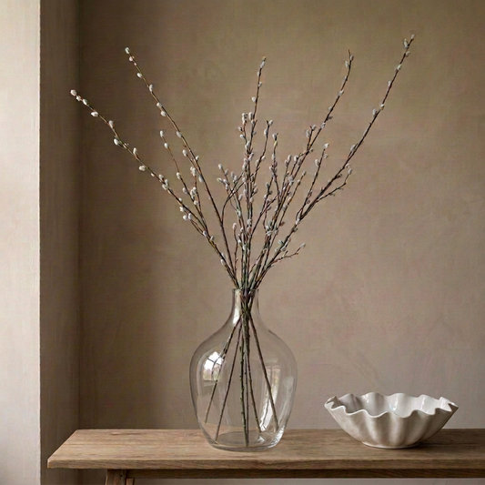 Clear glass vase with pussy willow branches on a wooden surface against a beige wall