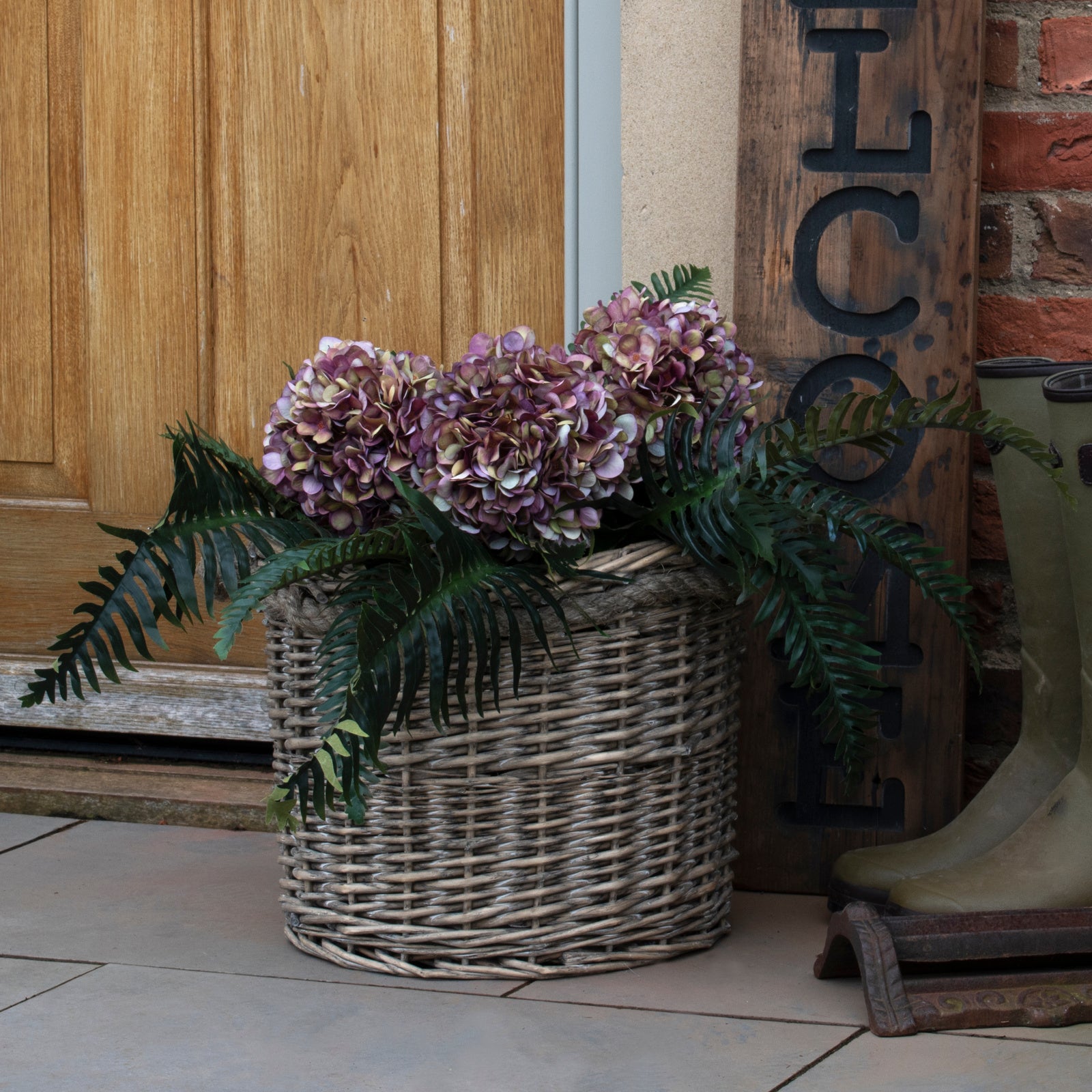 A natural woven wicker basket filled with purple hydrangea flowers and green fern leaves, placed on a tiled floor in front of a wooden door and brick wall.