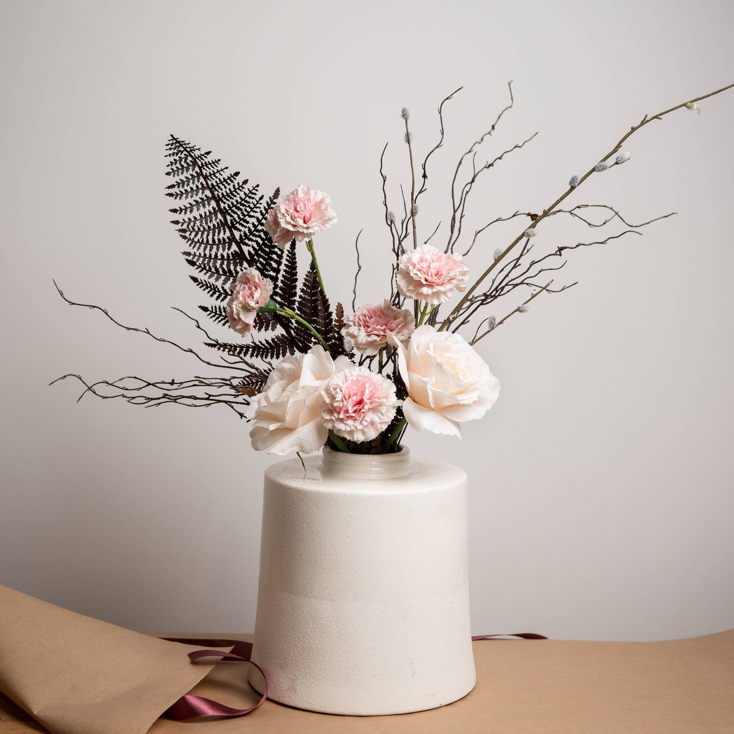 A white cylindrical vase holding a floral arrangement with pale pink and white flowers, black fern leaves, and thin bare branches, set on a tan surface against a light gray wall.