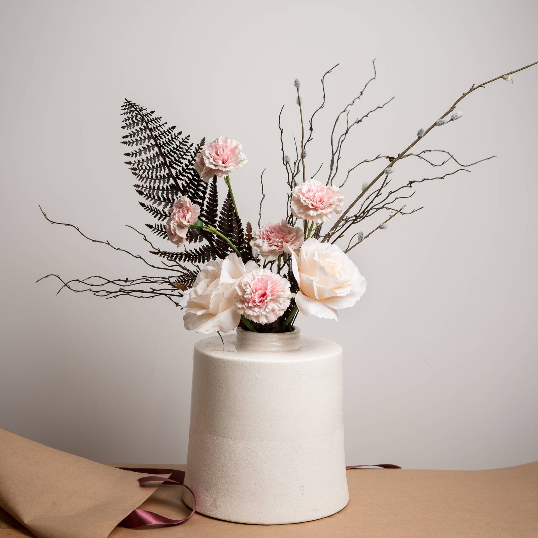 A white cylindrical vase holding a floral arrangement with pale pink and white flowers, black fern leaves, and thin bare branches, set on a tan surface against a light gray wall.