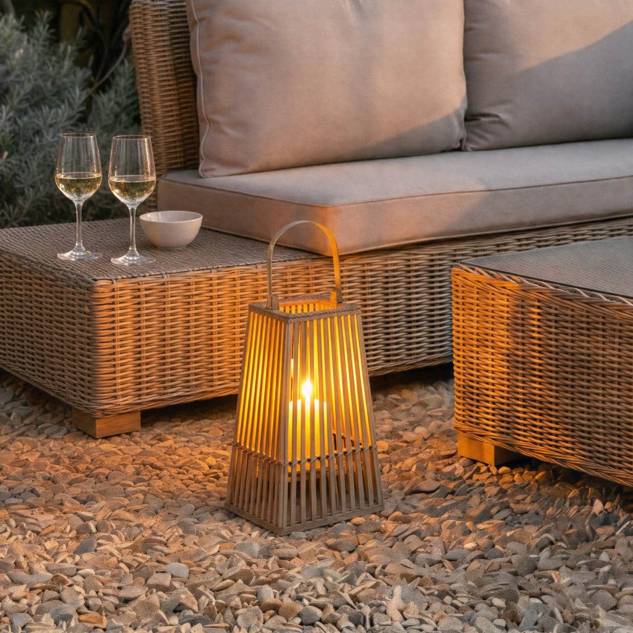 Small silo wicker outdoor or indoor lantern with glass candle holder glowing on a garden patio beside wicker garden furniture at dusk