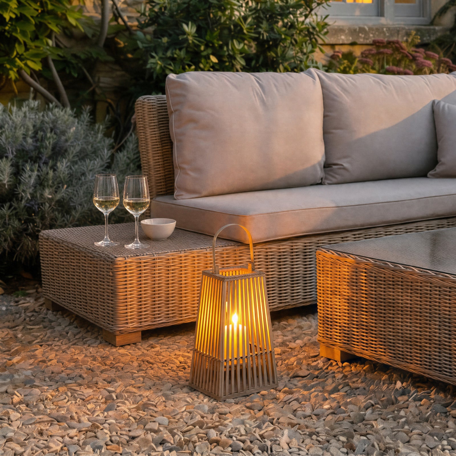 Outdoor garden patio setting with a sofa, coffee table, and wicker rattan lantern on a gravel surface.