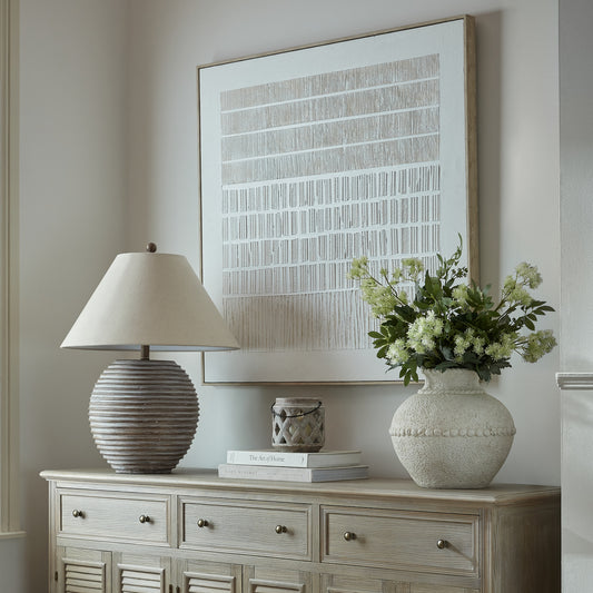 Large square neutral textured art line canvas in a slim natural oak frame, displayed above a light weathered oak sideboard styled with a ribbed ceramic table lamp, a chalky white textured vase filled with faux green foliage and white floral stems, a small stack of coffee table books topped with a lattice-cut lantern, against a warm off-white wall.