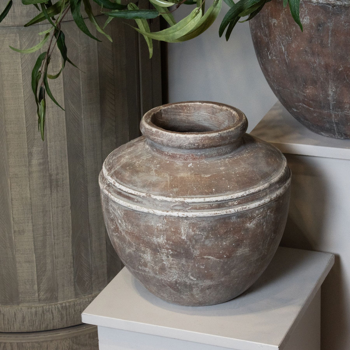 Decorative brown vase on a white surface with another vase and plant in the background.