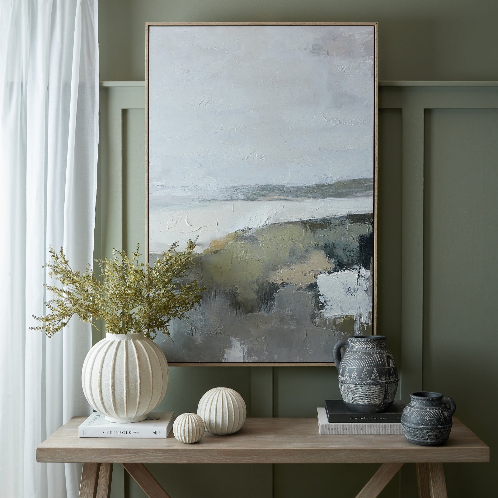 Decorative setup with abstract painting, vases, and books on a wooden table against a green wall.