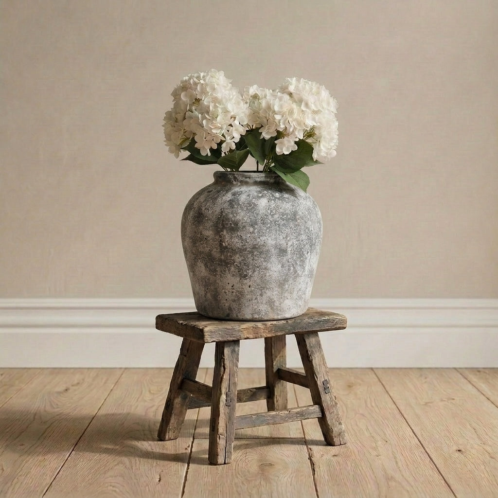Vintage-style vase with white flowers on a wooden stool against a beige wall.