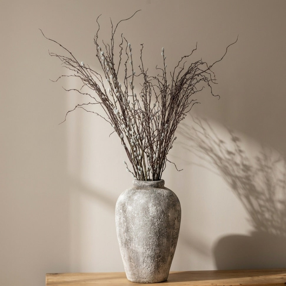 Decorative vase with artificial pussy willow and twig branches on a wooden surface against a beige wall
