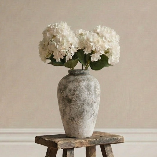 rustic stone aged-style vase with white flowers on a wooden stool against a beige wall