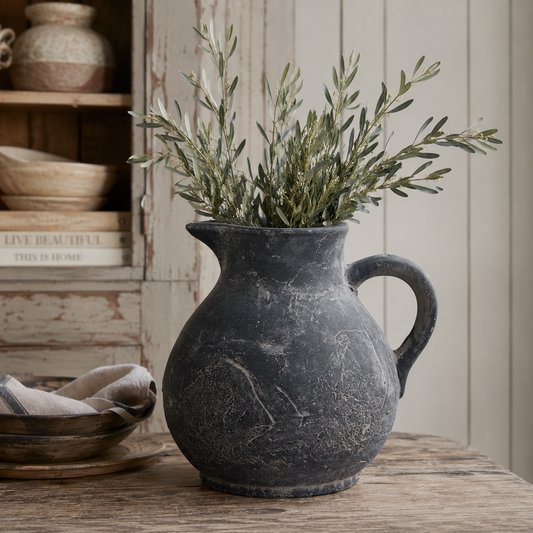 Amalfi grey ceramic jug with distressed stone finish styled on a reclaimed wooden surface with olive branches, beside a dark wooden bowl holding a striped linen cloth, against a white tongue-and-groove panelled wall