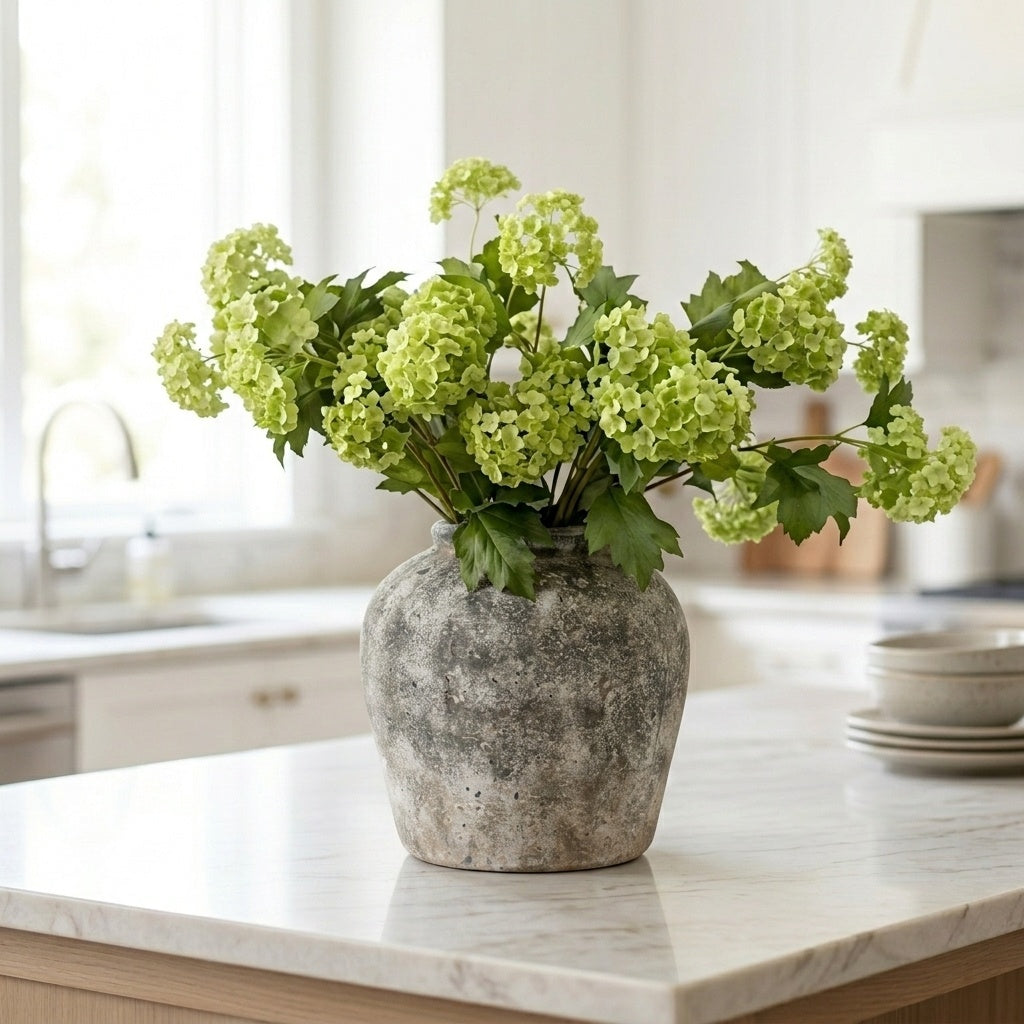 Green artificial Viburnum spray arranged in a rustic stone-effect vase on a white marble kitchen island in a bright Hamptons-style kitchen
