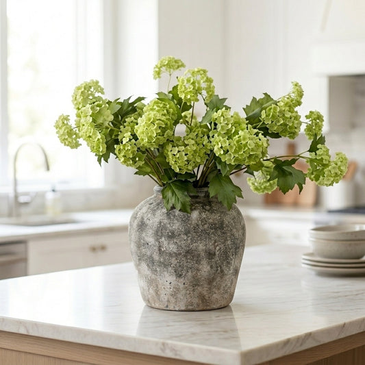 Green artificial Viburnum spray arranged in a rustic stone-effect vase on a white marble kitchen island in a bright Hamptons-style kitchen