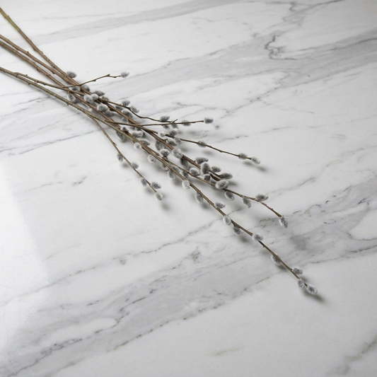 A decorative arrangement of bare brown  artificial pussy willow branches with small gray-white catkins, placed diagonally on a white marble surface with gray veining.