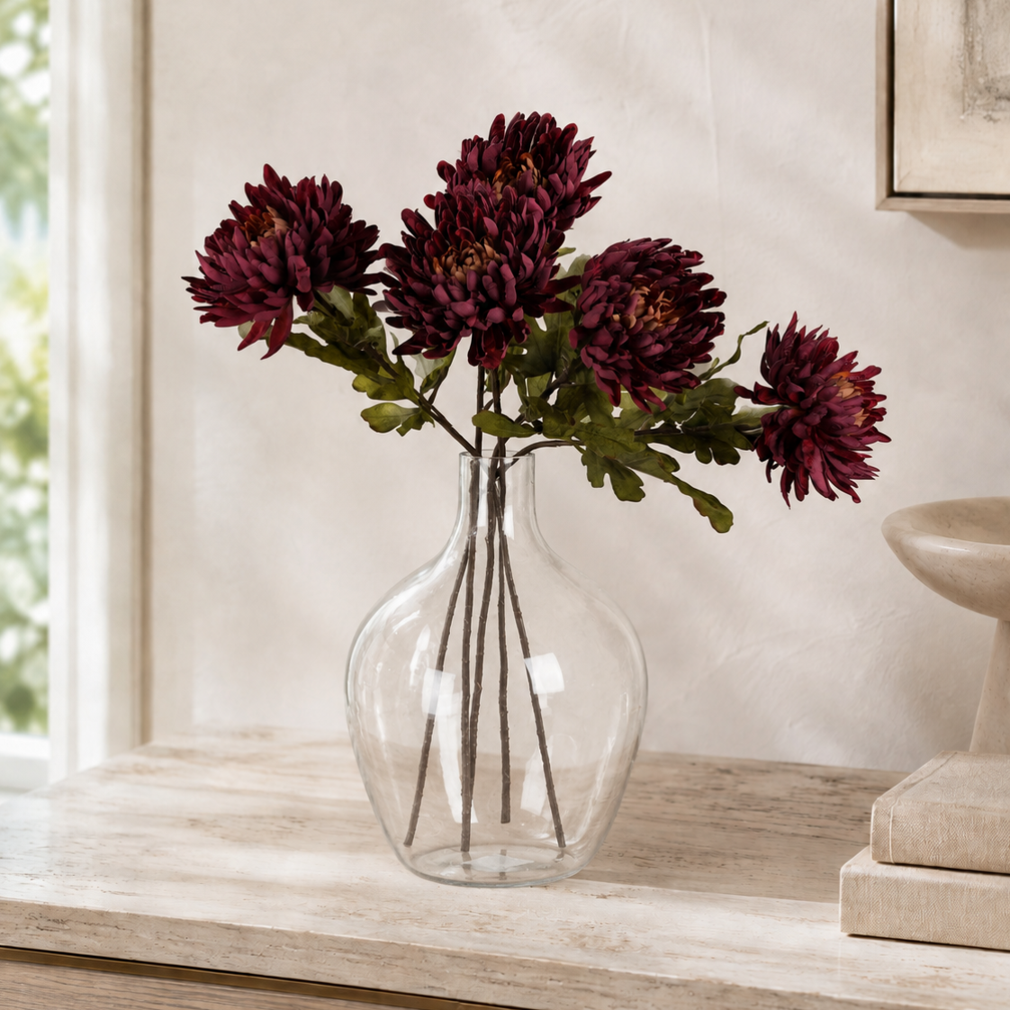 A clear glass vase filled with dark burgundy chrysanthemum flowers, styled on a light wood console table against a textured beige wall.