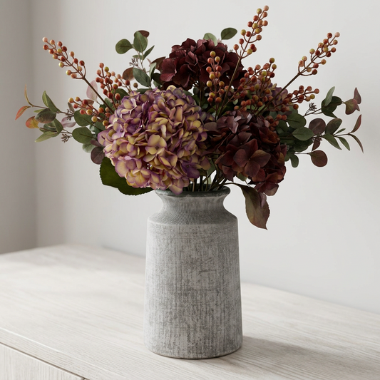 A faux floral arrangement in a textured gray ceramic vase, featuring purple and burgundy hydrangea blooms, dark berries, and green foliage, displayed on a light wood surface against a neutral wall.