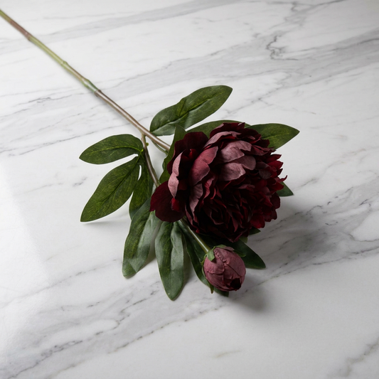 A single artificial peony stem with a deep burgundy bloom, green leaves, and a small bud, placed on a white marble surface.