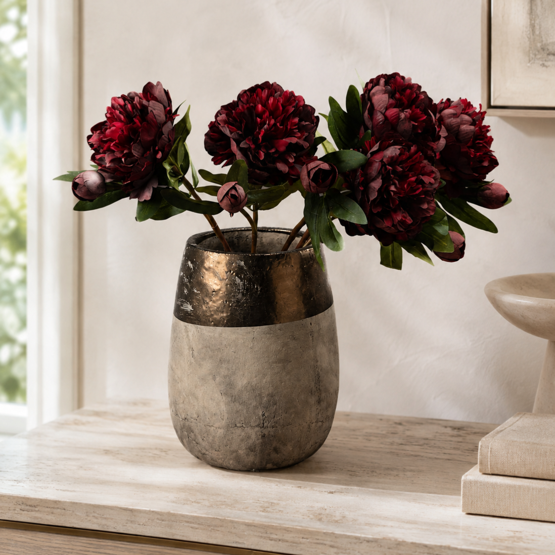 A decorative ceramic vase with a metallic gold top and a textured gray lower half, holding deep burgundy peony flowers with green leaves, set on a light wood console table against a neutral wall.