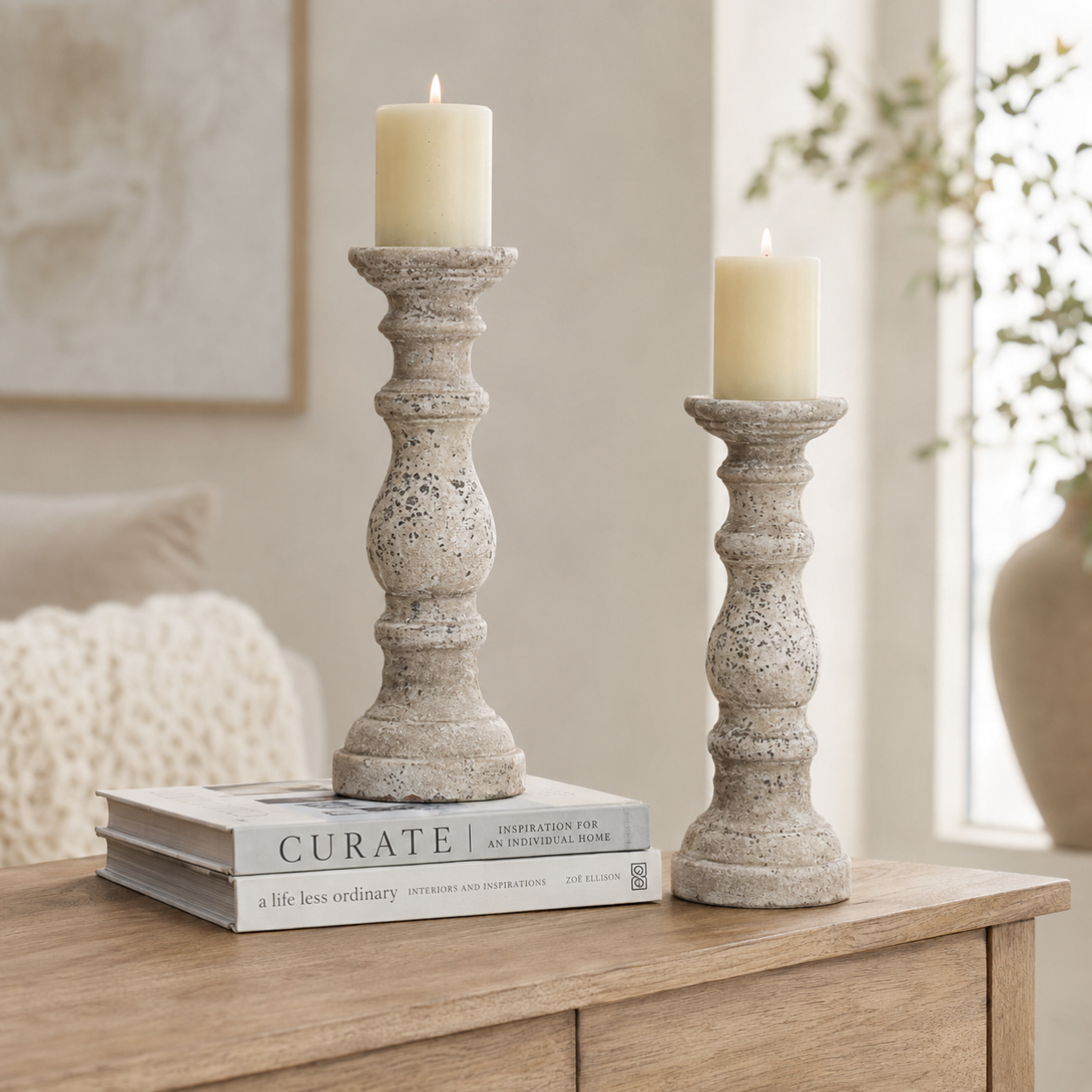 Two distressed stone-look candlestick holders with cream pillar candles, styled on a wooden console table with a book in a softly lit living room.