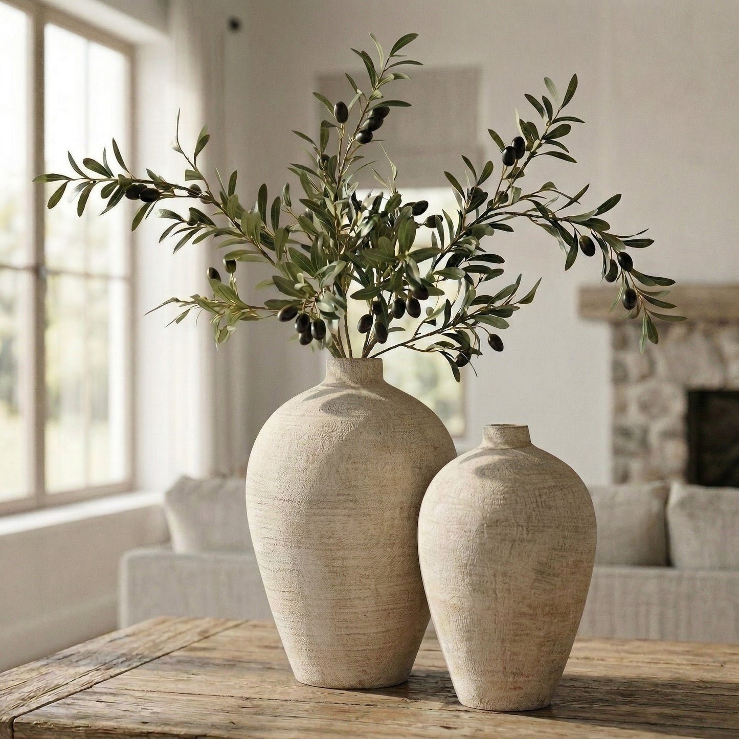 Two beige vases with artificial olive branches on a wooden table in a living room.