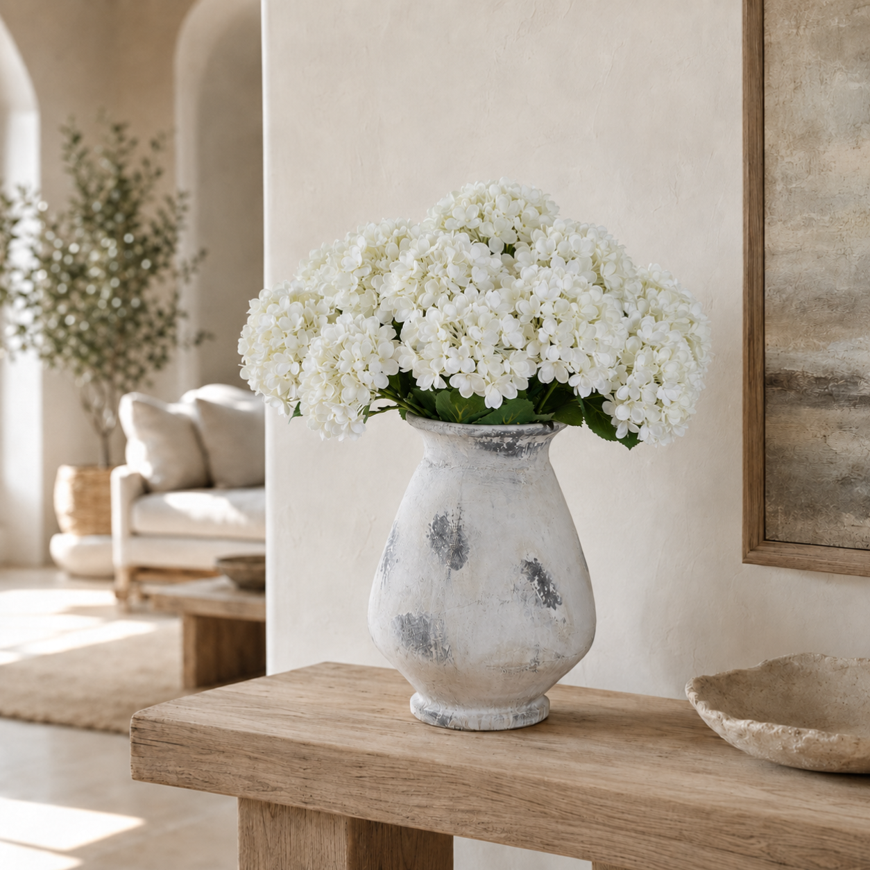 White hydrangea flowers arranged in a distressed white tall ceramic vase, placed on a wooden console table in a softly lit, neutral-toned interior room.