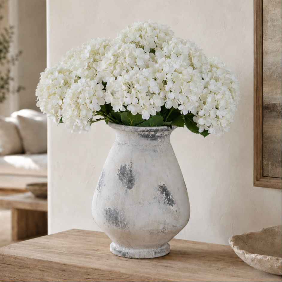 White hydrangea flowers arranged in a distressed white ceramic vase with gray mottled detailing, placed on a wooden table in a softly lit interior setting.