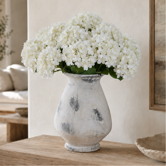 White hydrangea flowers arranged in a distressed white ceramic vase with gray mottled detailing, placed on a wooden table in a softly lit interior setting.