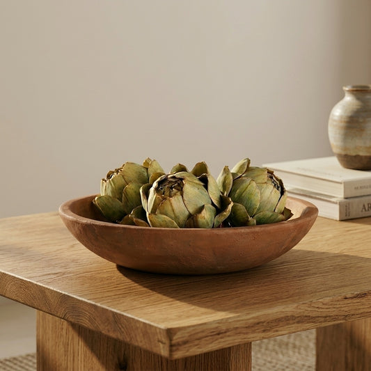 Three faux decorative artichokes styled as a bowl filler inside a wide shallow terracotta bowl on a solid oak coffee table, with stacked cream books and a stone ceramic vase in the background against a warm grey wall