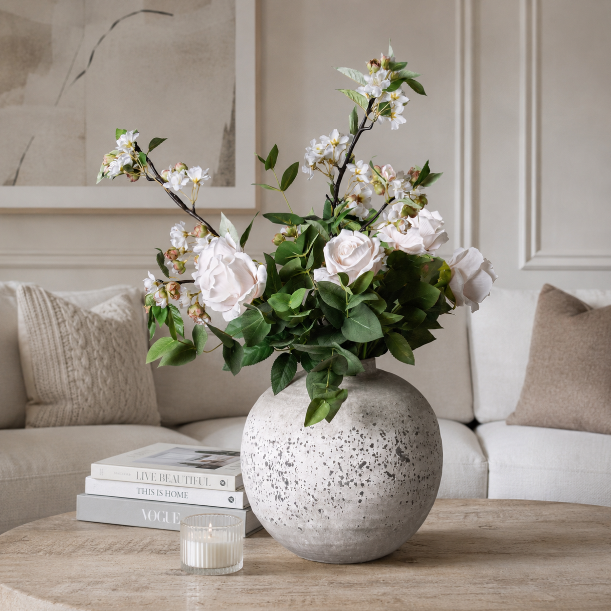 Large spherical grey stone-effect ceramic vase styled on a wood-grain coffee table with blush roses and white blossom stems, beside stacked books and a glass candle votive
