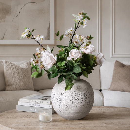 Large spherical grey stone-effect ceramic vase styled on a wood-grain coffee table with blush roses and white blossom stems, beside stacked books and a glass candle votive